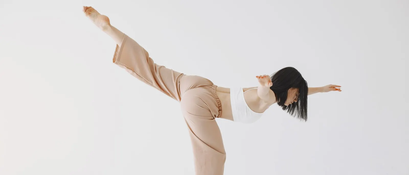 A woman balancing on one leg with the other leg raised high, against a plain white wall