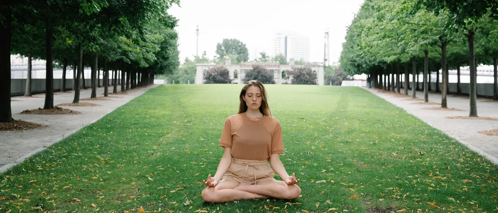 A woman meditating cross-legged on the grass of a tree-lined park